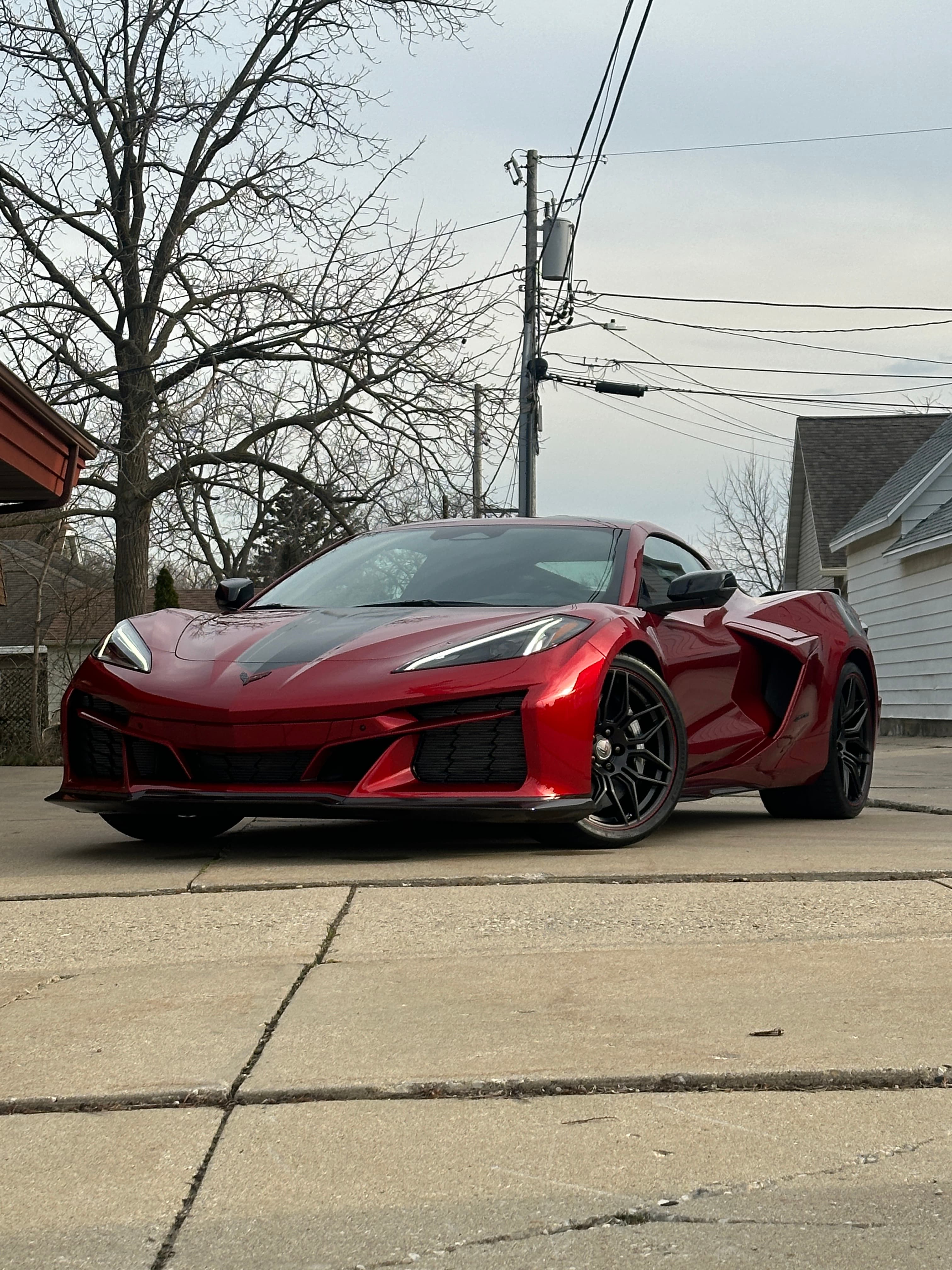 Metallic red Chevrolet Corvette C8 parked on a concrete driveway near a bare winter tree.
